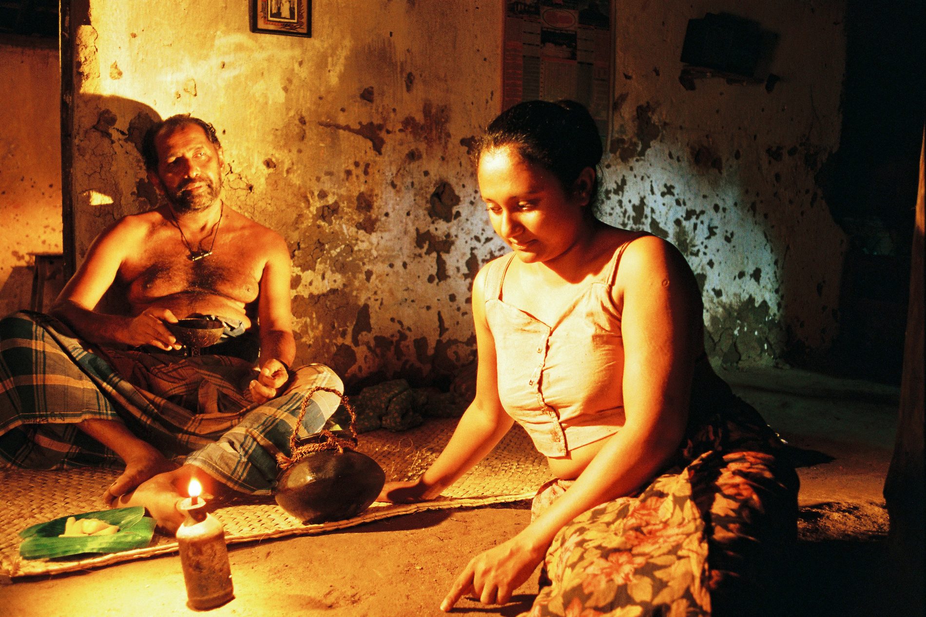Manoratne by lamplight in a mud-walled interior.